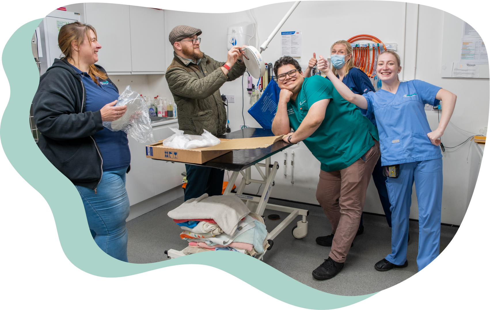 Five members of the Ashlands Vets team. They are standing in a veterinary consulting room, smiling and looking at the viewer. Four are wearing blue or green scrubs, the other is wearing a hat and a green padded jacket.